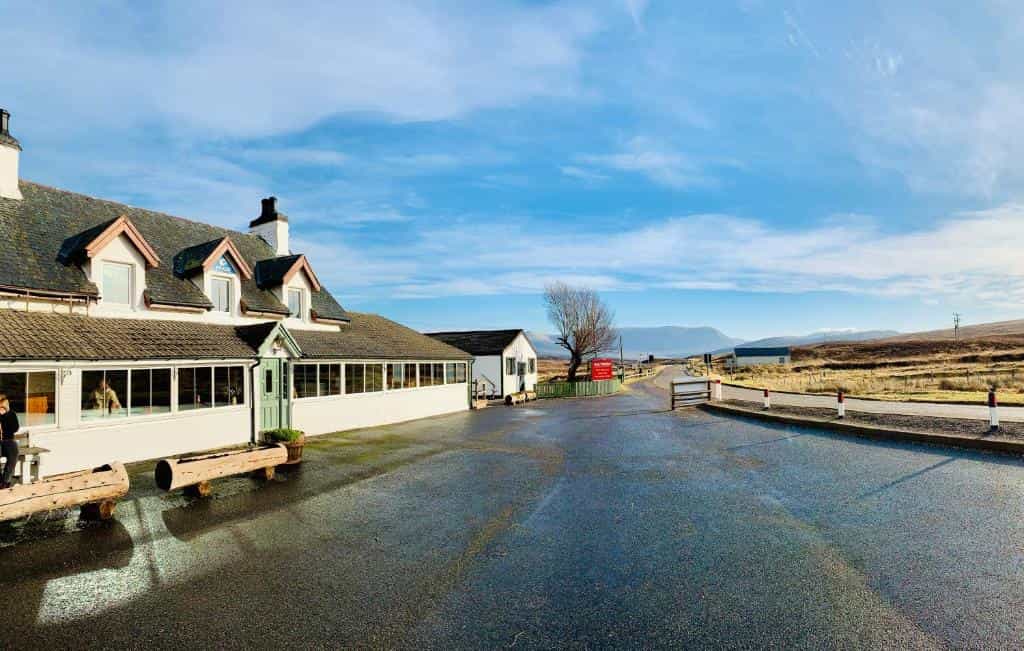 Aultguish Inn building with white walls and dark roof beside wet asphalt parking lot and distant hills.