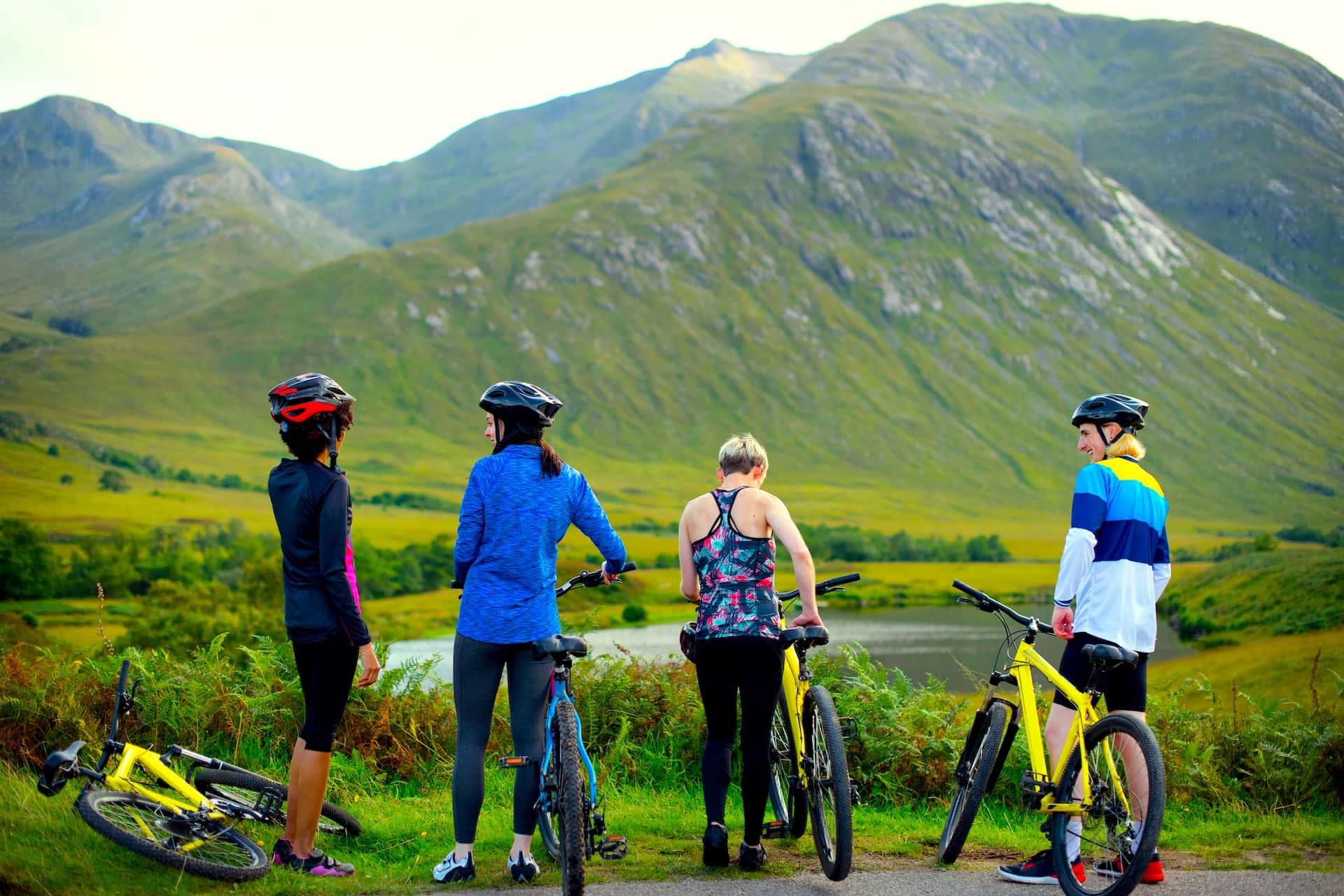 Cyclists with mountain bikes taking a break overlooking green mountains and a lake in the Highlands.