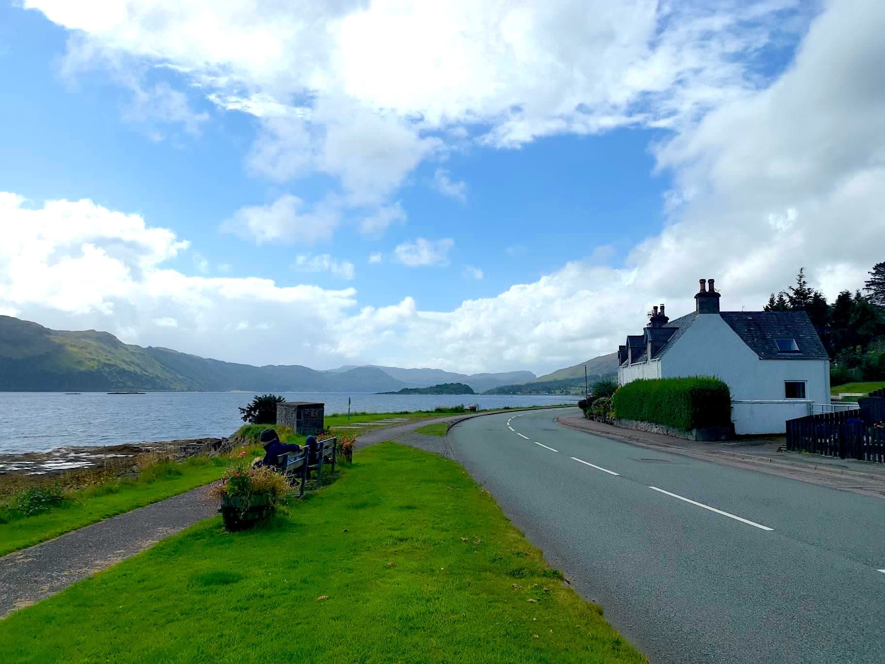 Roadside view of loch and mountains near white cottages, likely in the Scottish Highlands.