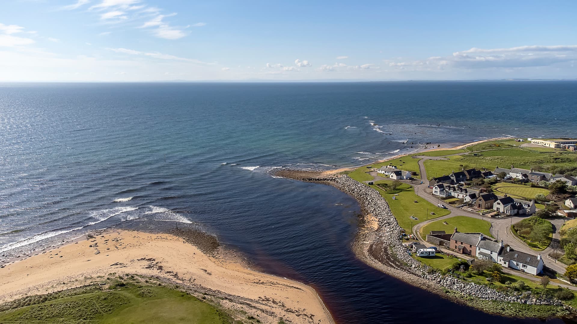 Aerial view of Brora coastline where a dark river meets the blue sea, with houses nearby.