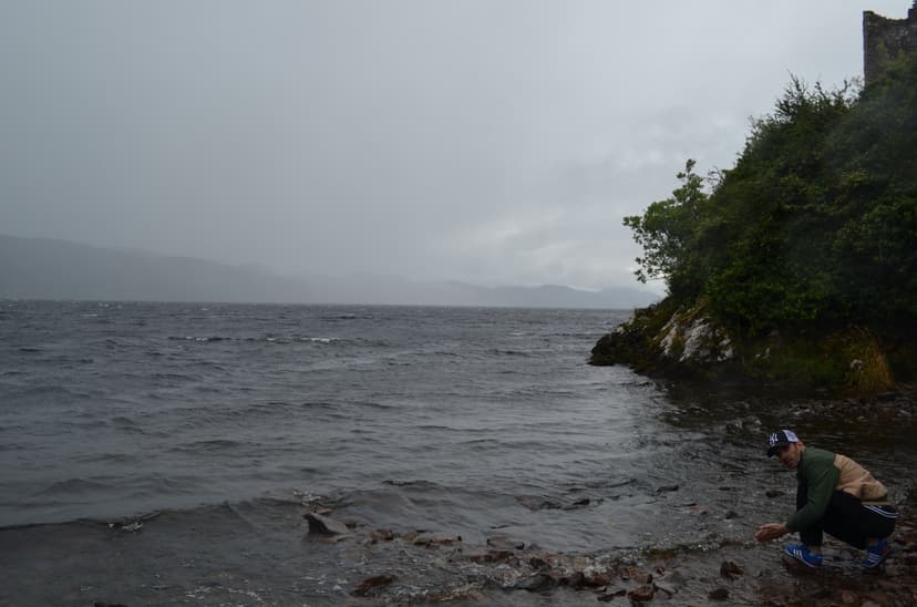 Person crouching by dark lake with overcast sky and distant mountains