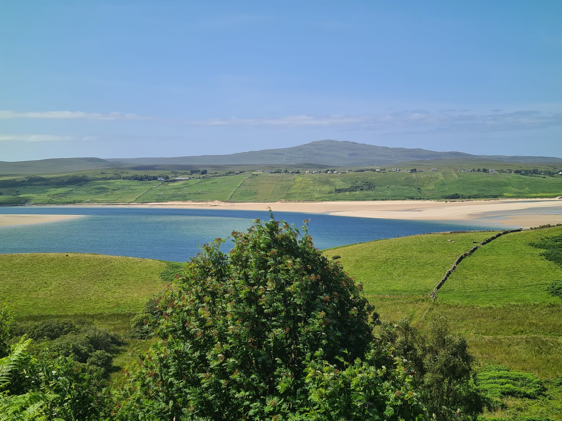Sandy estuary with blue water, green hills, and distant mountains under a blue sky