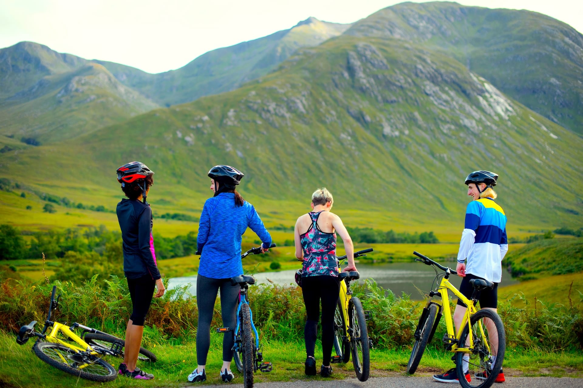 Cyclists with mountain bikes taking a break overlooking green mountains and a lake in the Highlands.