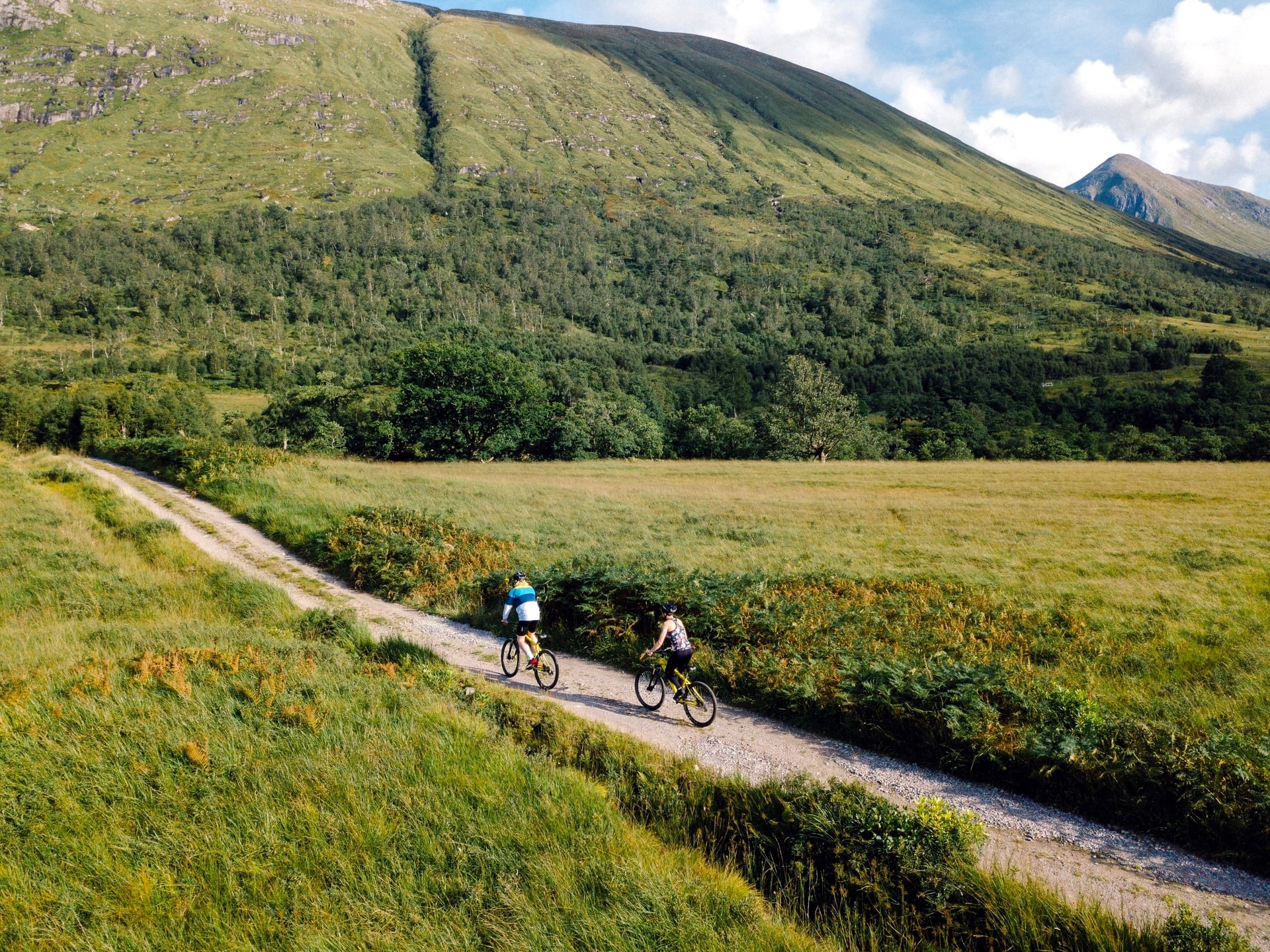 Couple cycling down a gravel road in the Scottish Highlands with green mountains.