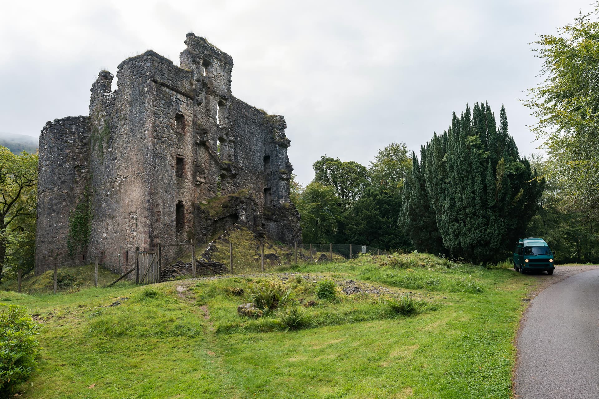 Ruins of Invergarry Castle beside a road with a green van and lush green trees.