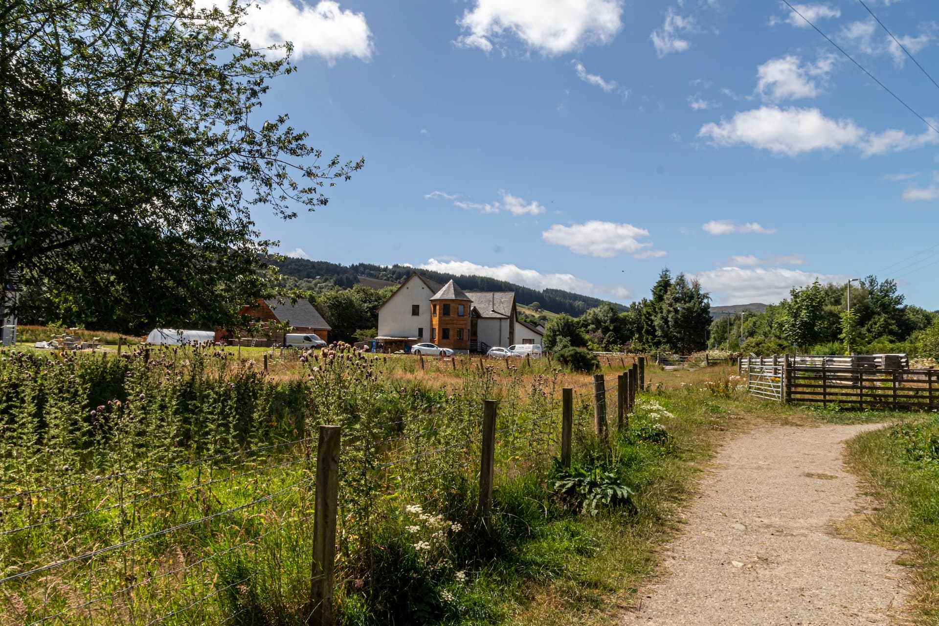 Dirt path leading past a wire fence through overgrown summer fields toward houses nestled in green hills near Drumnadrochit.