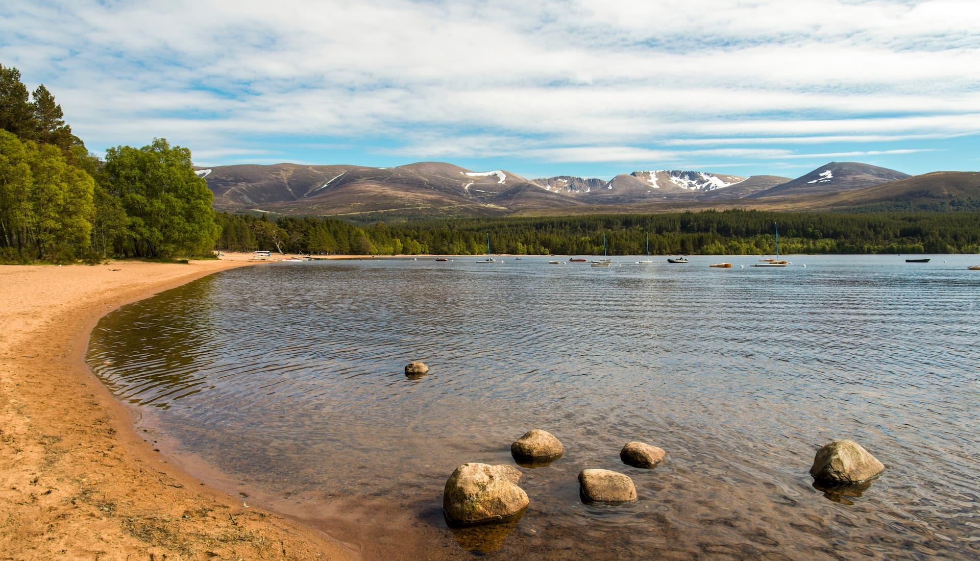 Sandy Loch Morlich shore with small boats, pine forest, and snow-dusted mountains.