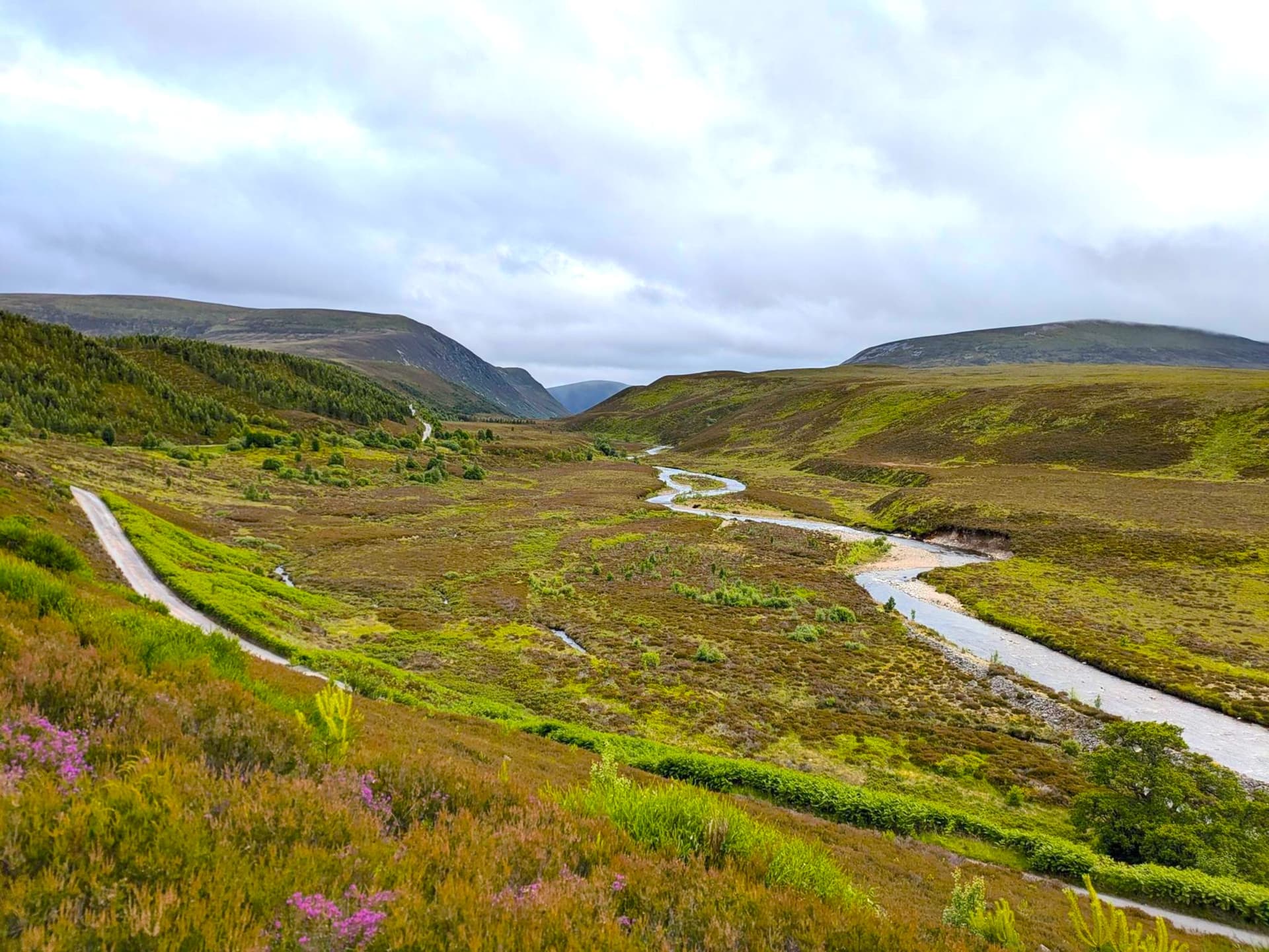 Winding river and road through green and heather-covered valley toward distant hills under cloudy sky near Bair Atholl.