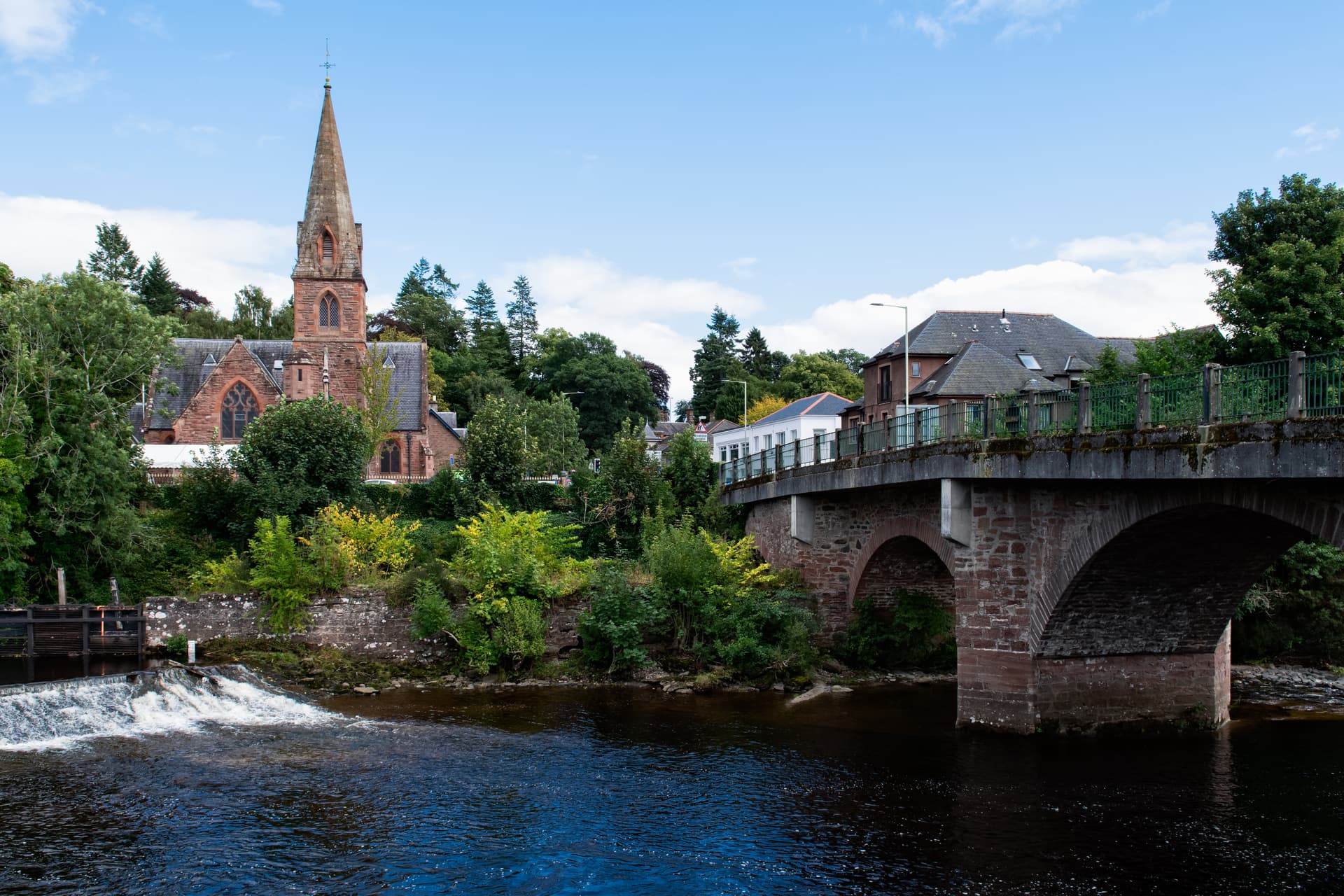 Stone church spire and arched bridge over dark river with small weir in Blairgowrie.