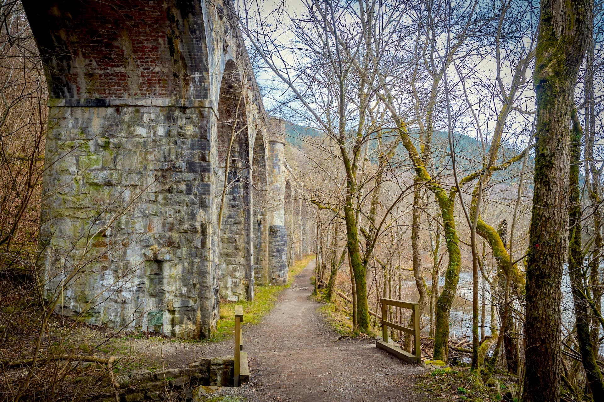 Stone viaduct arches tower over a dirt path beside a river, surrounded by bare trees in the Pass of Killiecrankie.
