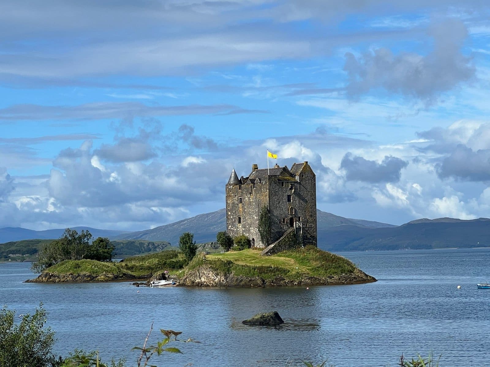 Stone castle on small island in water with mountains in background, yellow flag flying
